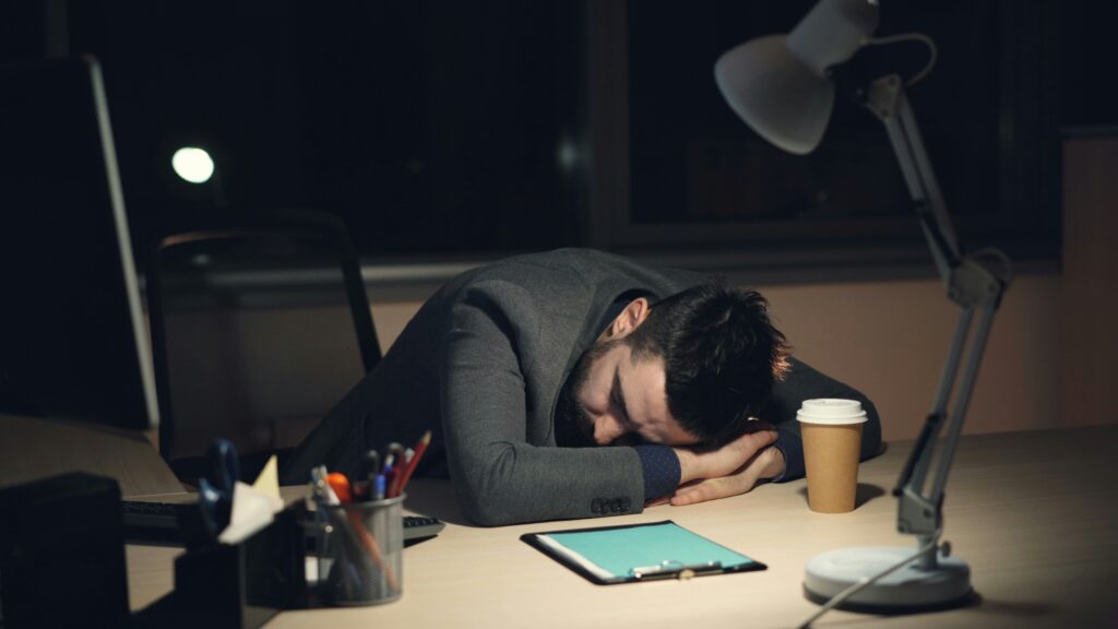 コーヒーとタブレットを持って机で寝ている男性。